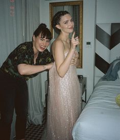 two women standing next to each other in front of a bed with a mirror on the wall