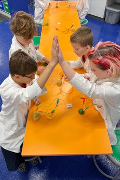 four children are sitting at a long table making something with wires and beads on it