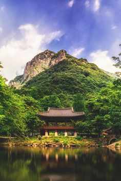 a small building sitting on top of a lush green hillside next to a lake in front of a mountain