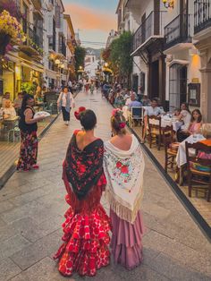 two women are walking down the street in front of some buildings with people sitting at tables