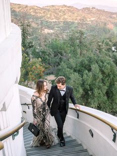 a man and woman are walking down the stairs in formal wear, with trees in the background