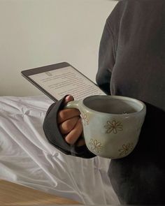 a person holding a coffee cup with a book in their hand while reading on the bed