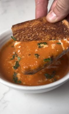 a grilled cheese sandwich being dipped into a bowl of tomato soup with parsley