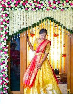 a woman in a yellow and red dress standing under a flower covered arch with flowers on it