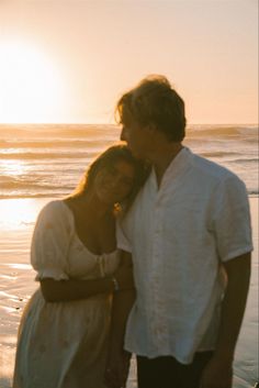 a man and woman standing next to each other on the beach near the ocean at sunset