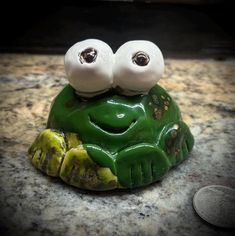 a green frog with two googly eyes sitting on top of a counter next to a coin