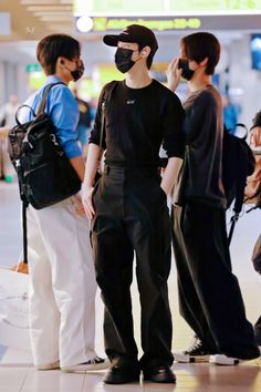 three people wearing masks and standing in an airport terminal, one is talking on his cell phone