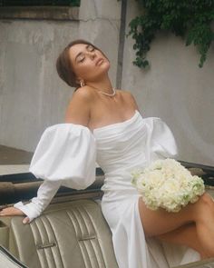 a woman in a white dress sitting on the back of a car holding a bouquet