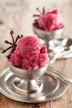 two silver bowls filled with red ice cream on top of a wooden table next to spoons