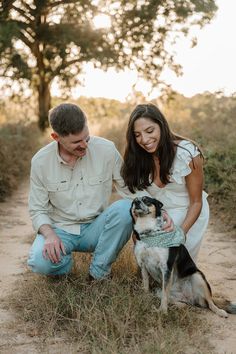 a man and woman sitting next to a dog on a dirt road with trees in the background