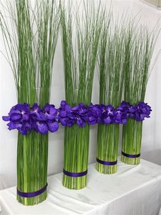 three tall vases with purple flowers and green grass in front of a white backdrop