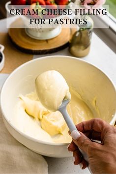 a hand holding a spoon with cream cheese filling for pastries in a white bowl