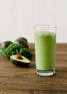 a glass filled with green smoothie next to an avocado on a wooden table