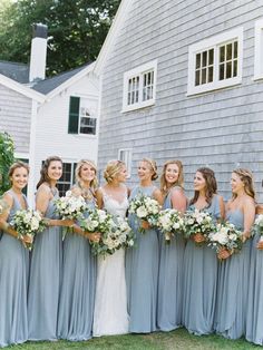 a group of women standing next to each other in front of a gray house holding bouquets