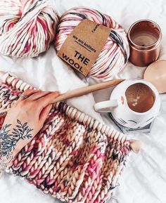 a woman's hand holding a cup of coffee next to some yarn and a wooden spoon