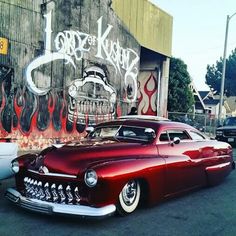 an old red car parked in front of a building with graffiti on the side of it