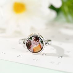 This photo ring is the best choice to make the memory eternal. Fashioned in sleek sterling silver, this ring features a charming daisy, and a shimmering round-cut stone is set at the center to complement the brilliance. With your own photo included, this beautiful piece will be more memorable. You can also wear it as your pendant to make your daisy-themed necklace dangling close to your heart.Carat Weight: 0.81 ctStone Size: 3,1.5,1.3 mmStone Type: Jeulia® StoneNumber of Stones: 28 Stone Shape: RoundStone Color: Citrine Yellow, Diamond WhiteWeight: 4.6 gWidth: 11.4 mmHeight: 5.9 mmThickness: 3.1 mmMaterial: 925 SilverPlating Color: Silver, Rose Gold Classic Round Birth Flower Jewelry, Silver Rings For Mother's Day, Round Jewelry With Polished Finish For Mother's Day, Elegant White Gold Round Pendant Ring, White Gold Flower Ring With Birthstone, Rose Gold Round Ring With Birth Flower Detail, Rose Gold Round Ring With Birth Flower, Personalized Silver Flower Ring For Anniversary, Anniversary Diamond Ring With Bezel Setting