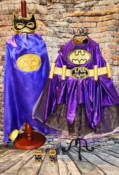 two purple and gold costumes on display in front of a brick wall, one wearing a batman cape