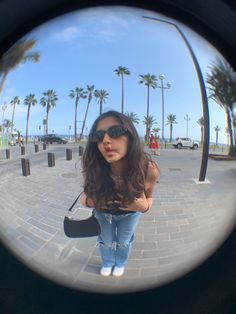 the reflection of a woman wearing sunglasses is seen through a circular lens in front of palm trees