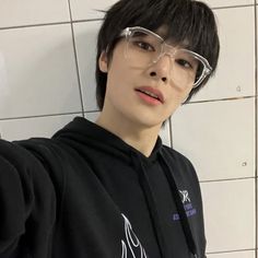 a young man wearing clear glasses in front of a white tile wall with his hand on his hip