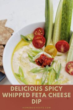 a white bowl filled with dip surrounded by sliced cucumbers and tomatoes, next to crackers