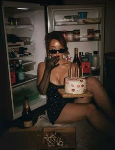 a woman sitting in front of an open refrigerator holding a cake with candles on it