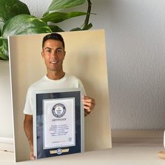 a man holding up a framed award in front of a potted plant and white wall