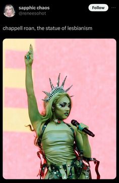 a woman with green makeup and headdress holding a microphone in front of a pink background