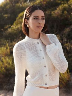 a woman wearing a white sweater and skirt standing in front of some bushes with her hand on her chin