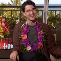 a man sitting down wearing a lei with flowers around his neck and smiling at the camera