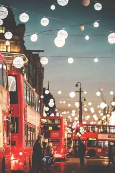 red double decker buses driving down a street with christmas lights hanging from the wires above