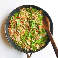 a skillet filled with rice and vegetables on top of a white table next to a wooden spoon