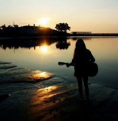 a woman is standing on the beach with her guitar and looking out at the water