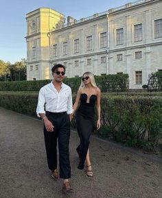 a man and woman walking in front of a large building with hedges on both sides