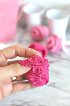 a hand holding a knitted pink bow on top of a marble table next to cups and saucers