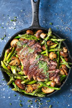 a skillet filled with asparagus, mushrooms and steak on a blue surface