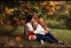 a mother and her child sitting under a tree in the fall time with leaves all around them