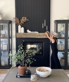 a woman in a black dress and hat standing next to a fireplace