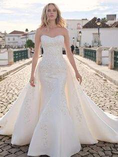 a woman in a white wedding dress standing on a cobblestone road with her arms behind her back