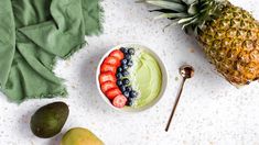 a bowl of fruit is sitting next to two pineapples and an avocado