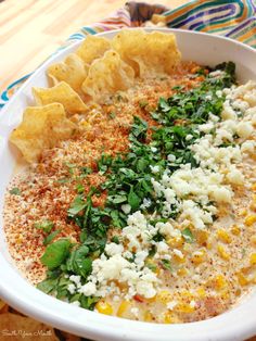 a white bowl filled with corn, cheese and herbs next to tortilla chips