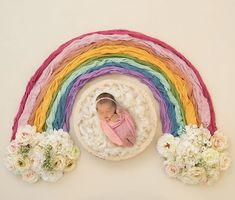 a baby is laying on top of a rainbow with flowers in the foreground and a white wall behind it