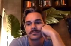 a man with a moustache on his face is taking a selfie in front of a bookshelf