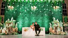 a bride and groom sitting on a couch in front of a wall with greenery