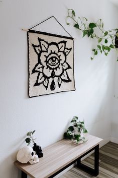 a wall hanging on the side of a white wall next to a wooden table with plants