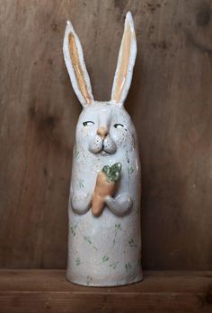 a ceramic rabbit holding a carrot on top of a wooden shelf next to a wall