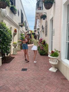 two women are walking down an alley way with potted plants on either side of them