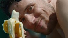 a shirtless man holding a peeled banana in his right hand and looking at the camera