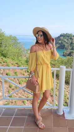 a woman in a yellow dress and hat standing on a balcony with her handbag