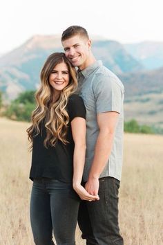 a man and woman are standing together in the grass with mountains in the back ground